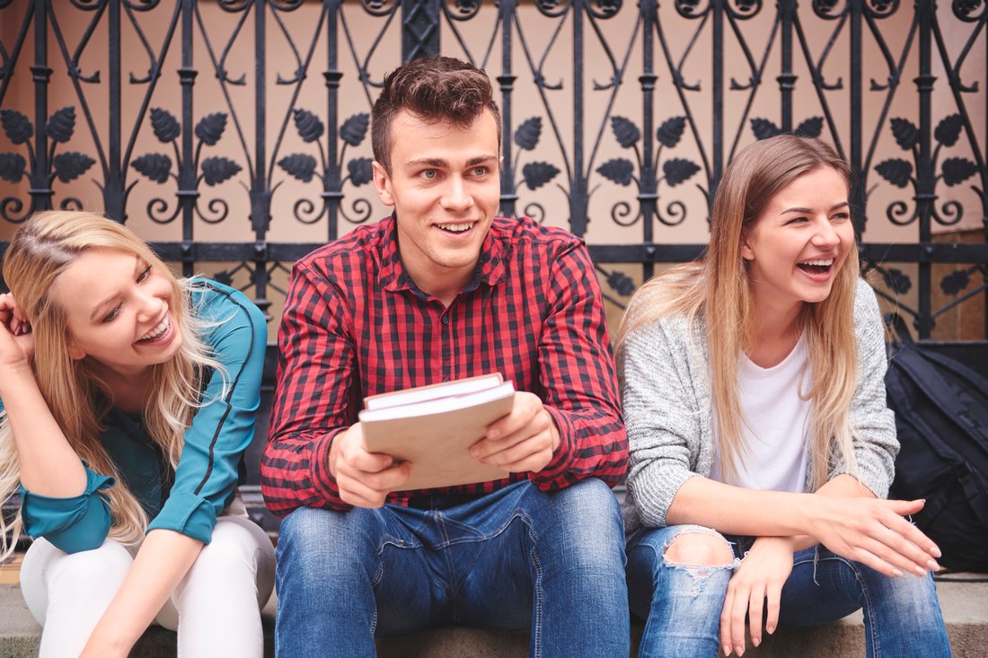Troje roześmianych studentów z książkami siedzi razem przed budynkiem uczelni